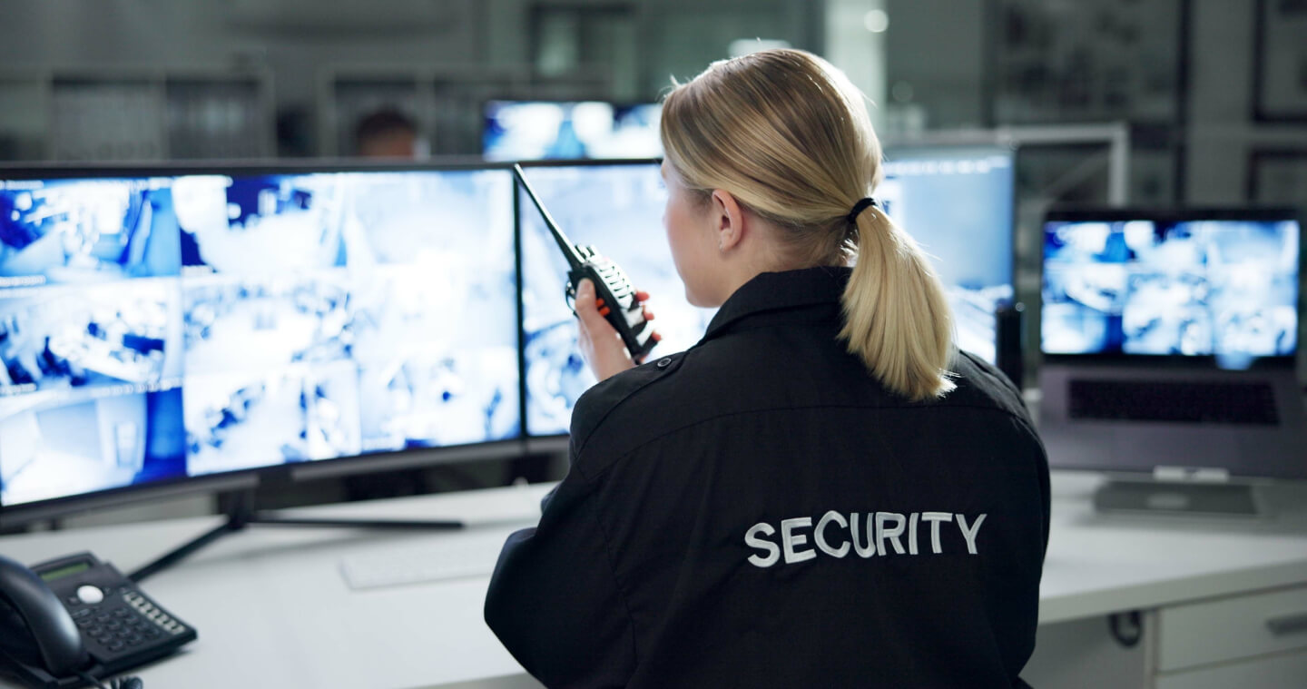 A woman sitting in front of monitors looking at security footage while talking through a hand help radio.